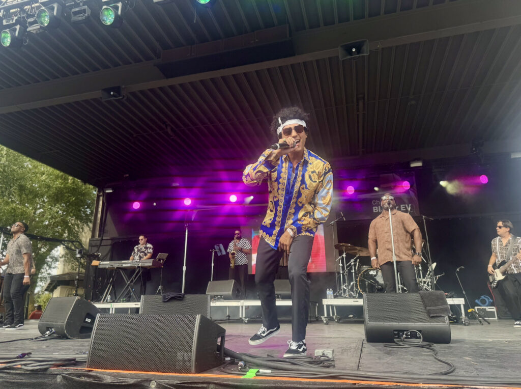 The lead singer of 24K Magic Band wears a blue and gold shirt as he leans forward on stage and sings into his microphone. 24K Magic is a Bruno Mars tribute band and performed at the New York State Fair on September 1, 2025.