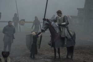 In "A Knight of the Seven Kingdoms" a knight sits unmasked on his horse in chainmail as his squire, a small, bald boy hands him his lance.