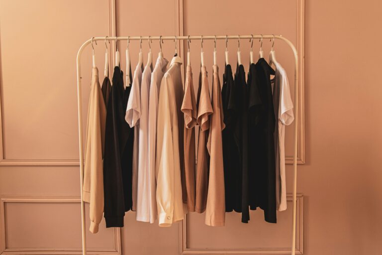 A collection of neutral colored blazers and shirts on a light-colored coat rack in front of a white wall.