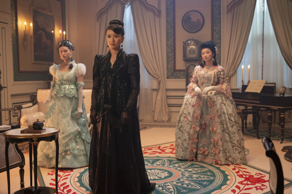 In Bridgerton Season 4, a woman in a black dress stands in the middle of two younger women in colorful, regency gowns in an elaborate sitting room.