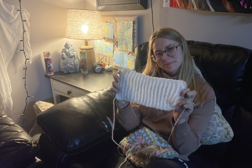 In a living room, a blonde woman sits in a black leather chair and proudly holds up a square of whit fabric she is crocheting.