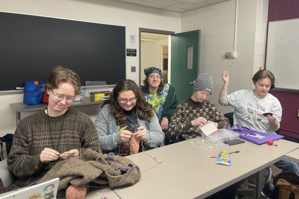 In a classroom, a group of students sit at a white table, all working on knitting and crocheting with colorful yarn.