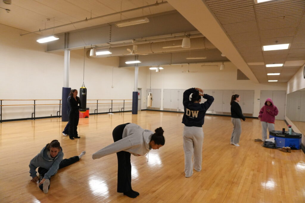 A group of students dressed in sweats stand in a dance studio in front of a mirror and stretch before rehearsal.