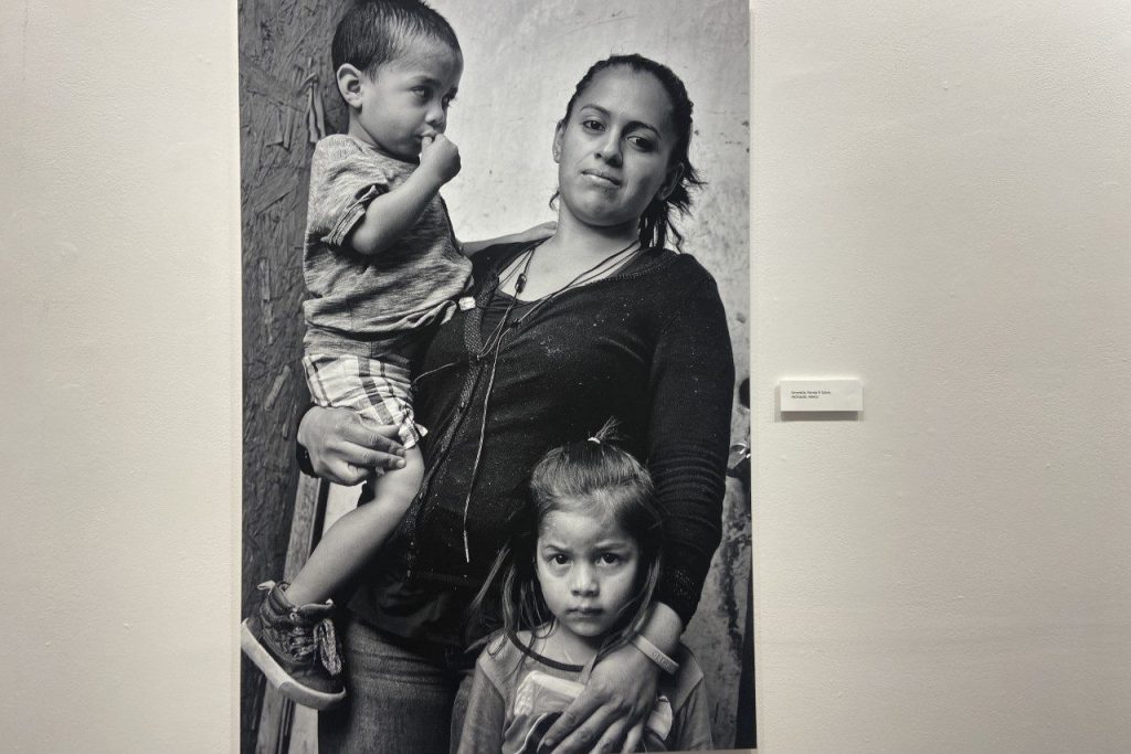 Woman with her toddler-age son on her right hip and her left hand draped over her small daughter holding her close. All looking at the camera.