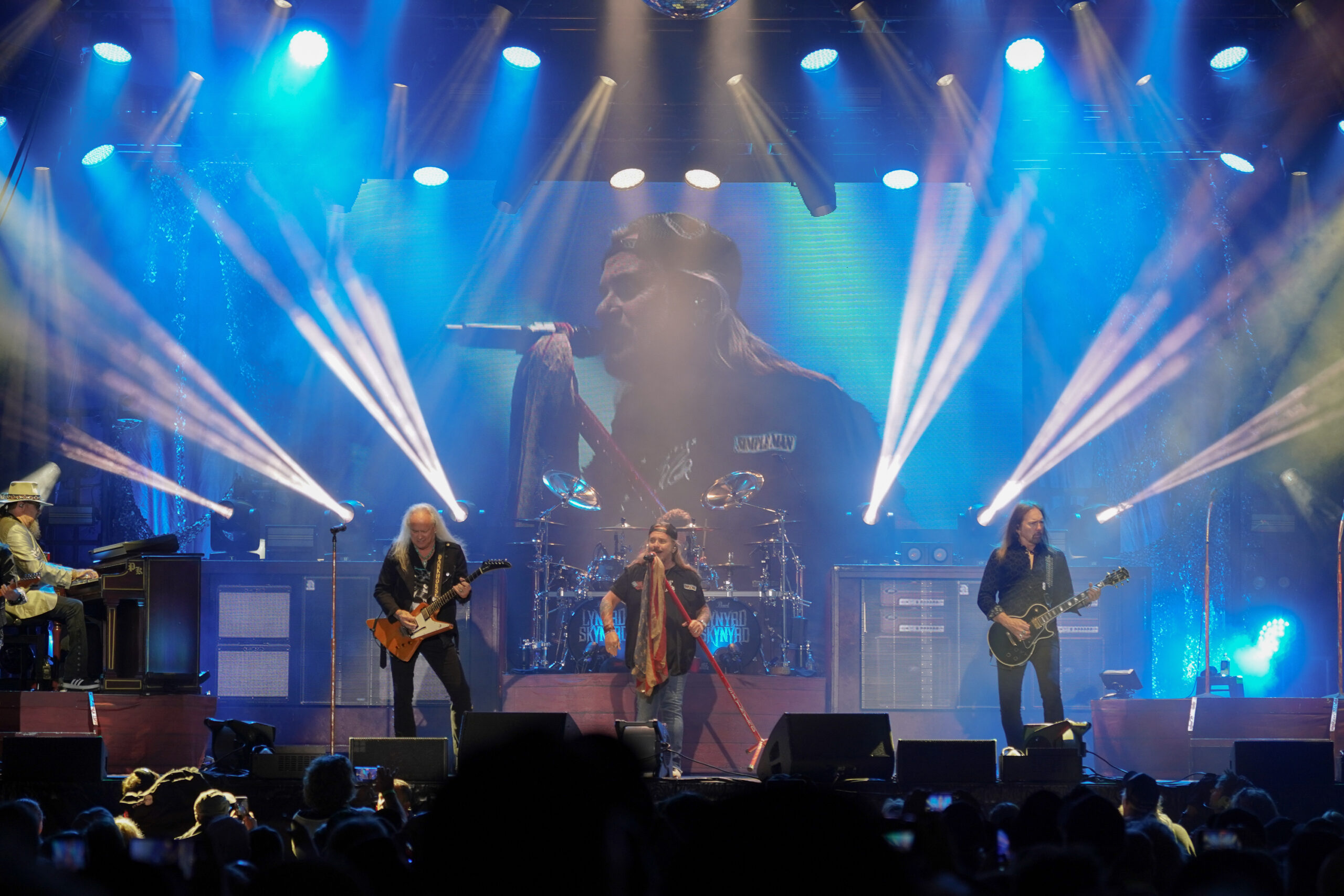 Lynyrd Skynyrd performs in front of a large crowd with a blue background and white lights