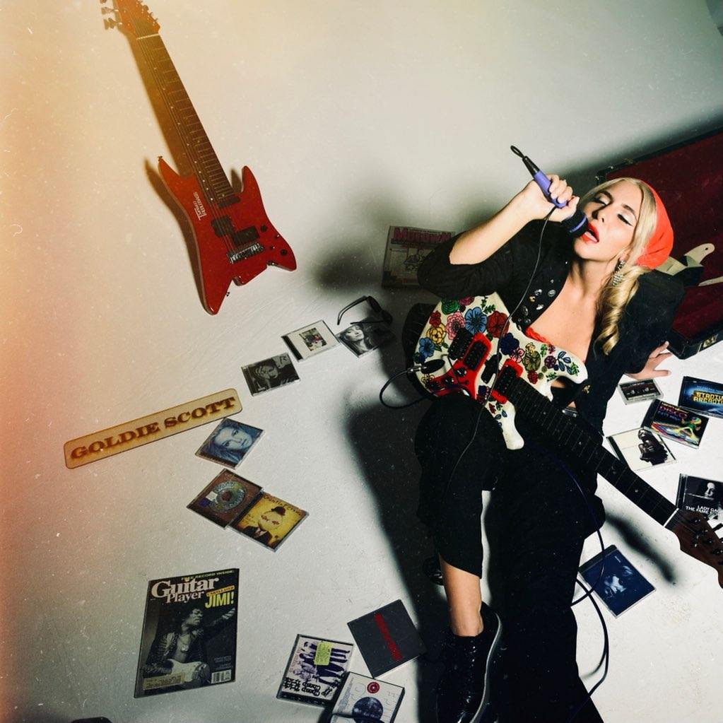 Goldie Scott singing into a microphone surrounded by CDs and guitars