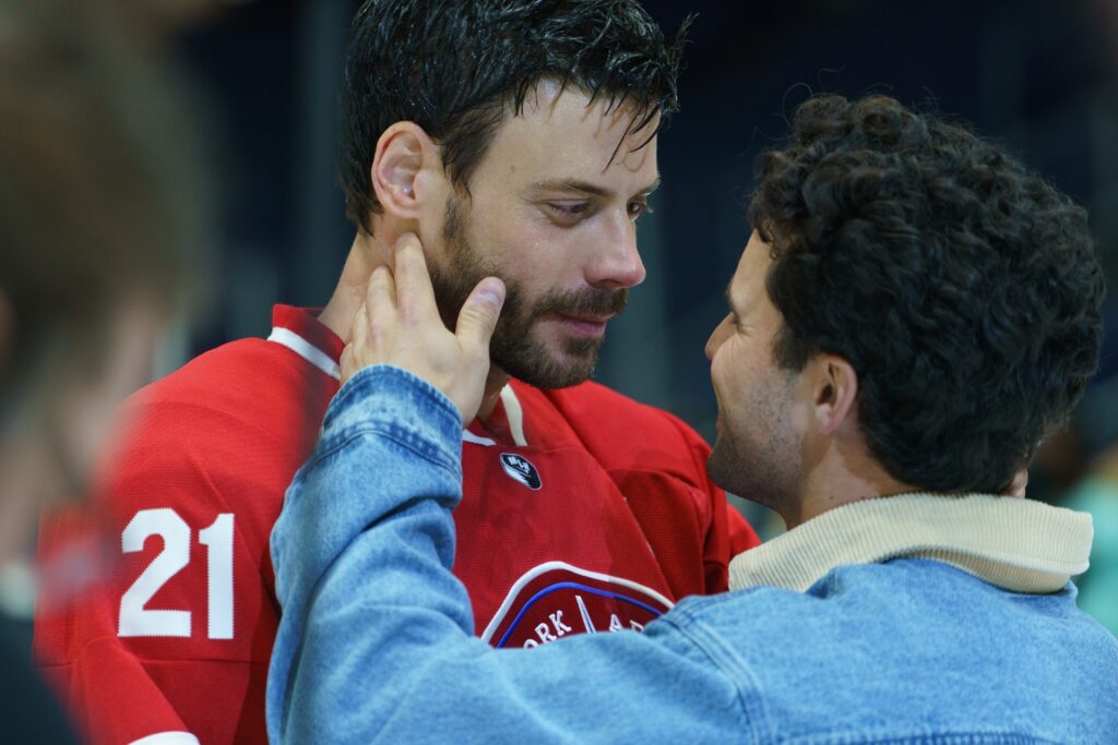 A male hockey player in a red jersey looks lovingly at a man in a jean jacket as they hold each other close.