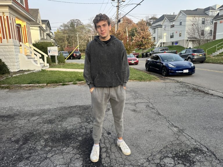 A man stands in the driveway of a house dressed in a dark sweatshirt and grey sweatpants.