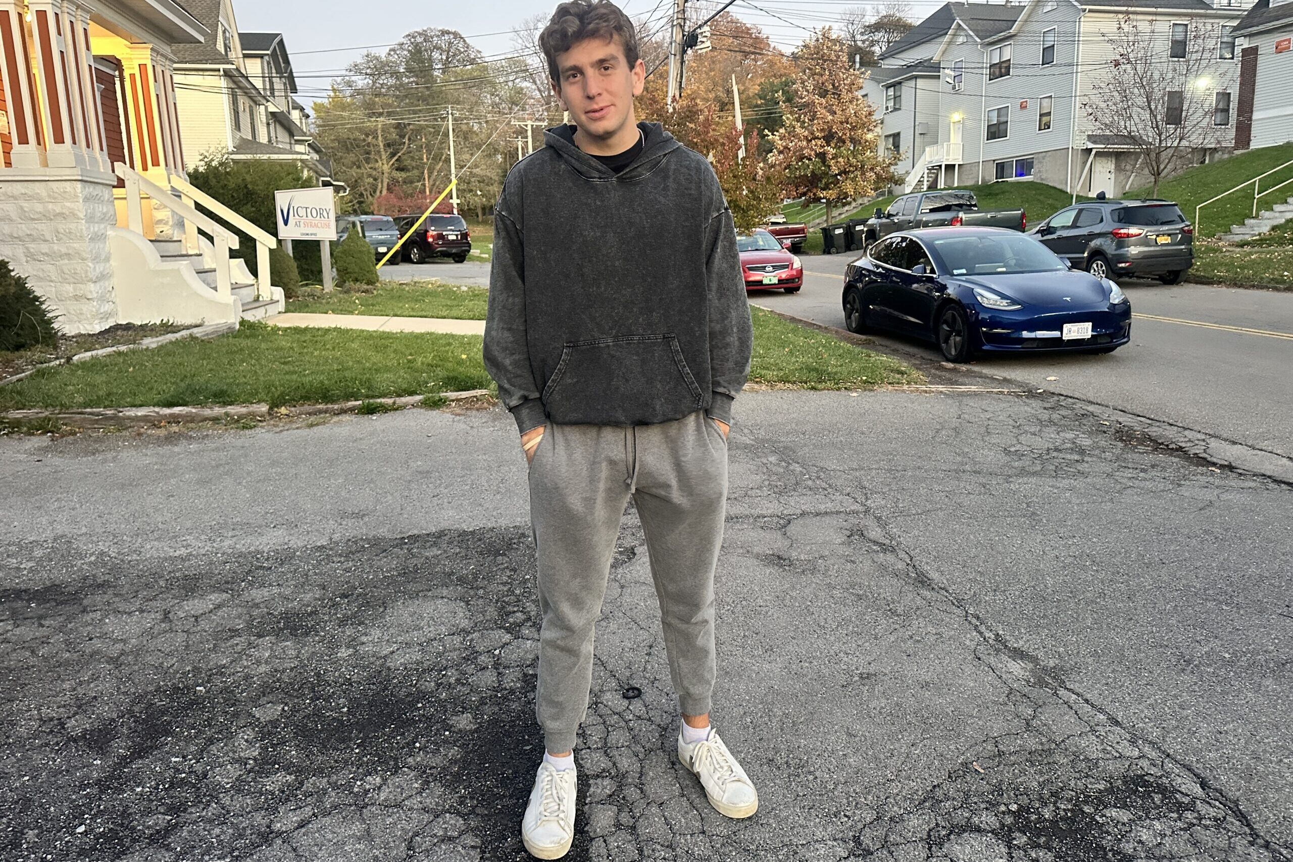 A man stands in the driveway of a house dressed in a dark sweatshirt and grey sweatpants.