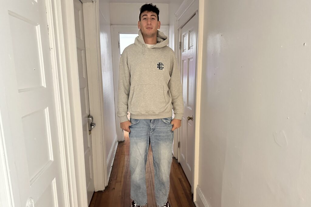 A man stands in a hallway dressed in a grey hoodie and light wash jeans.
