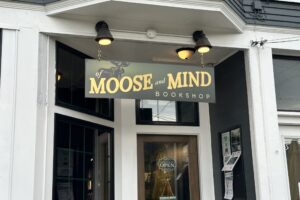 A green sign with the words "Of Moose and Mind" written in yellow text hangs white store with large glass windows.