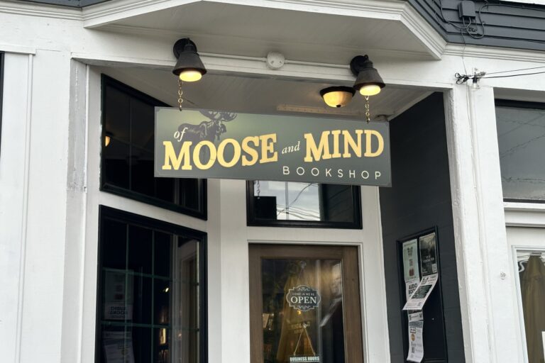 A green sign with the words "Of Moose and Mind" written in yellow text hangs white store with large glass windows.