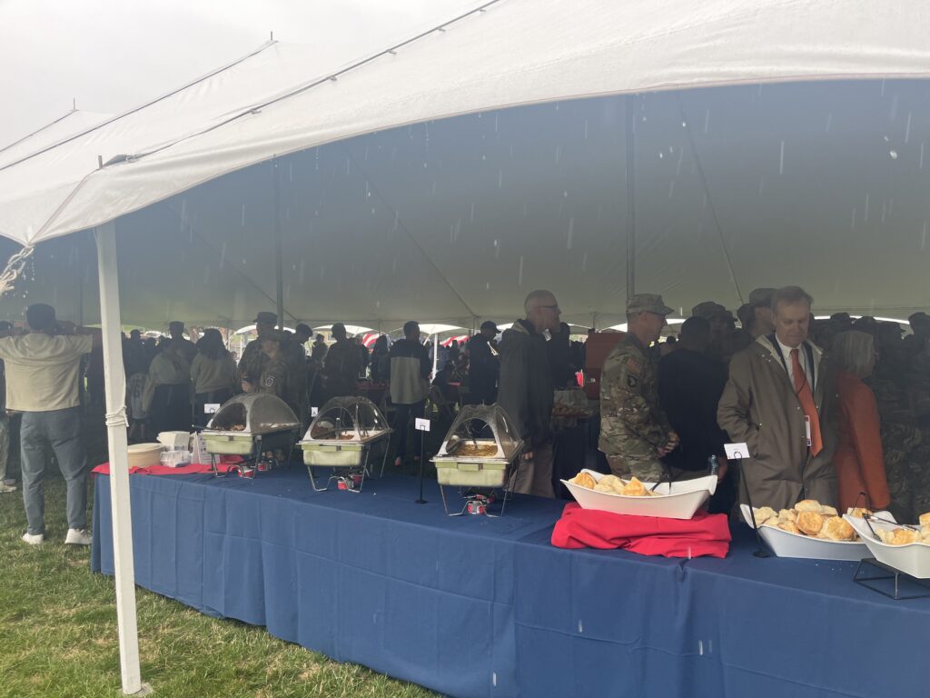 Food is served under a tent. A mix of people, some in military uniform, gather under the tent to speak.