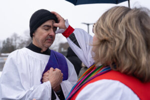 Father David in white robe with blue cloth receives ashes drawn into a cross shape by Mother DeDe in red robe.