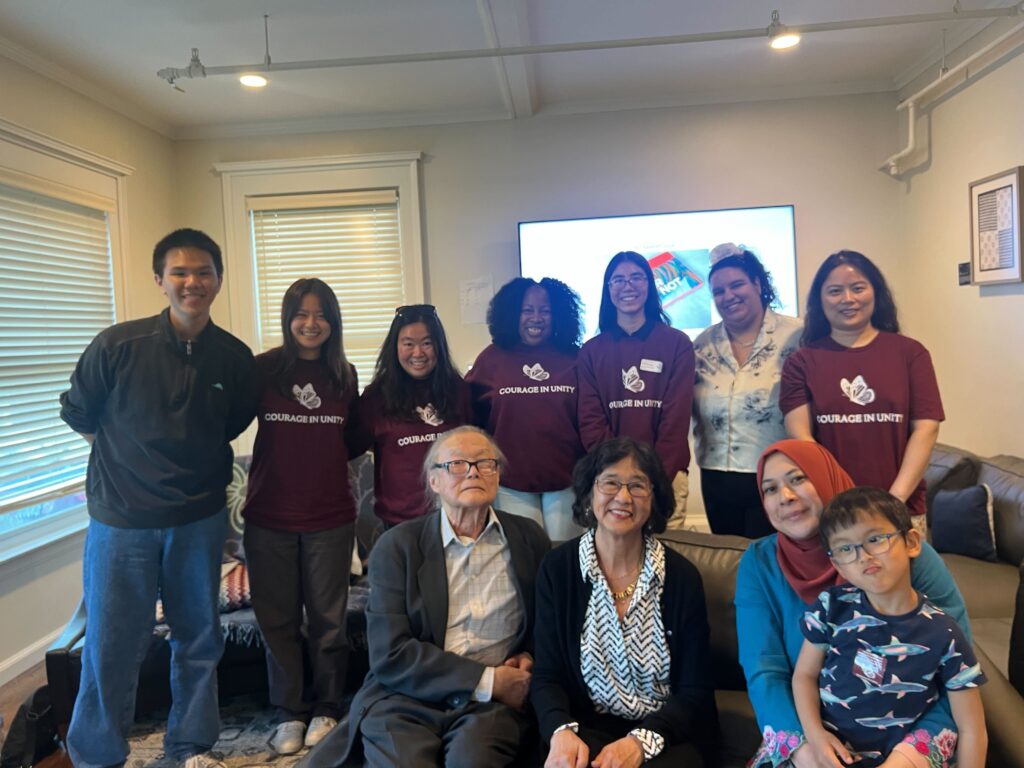 A group of young Asian American students stand behind professors sitting on a couch with a big TV and windows in the background.
