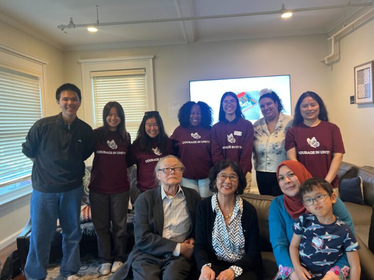 A group of young Asian American students stand behind professors sitting on a couch with a big TV and windows in the background.