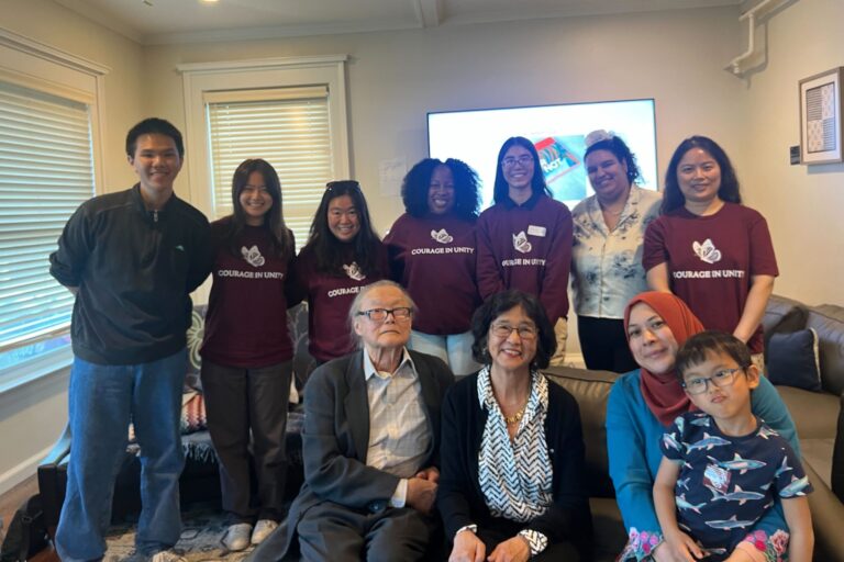 A group of young Asian American students stand behind professors sitting on a couch with a big TV and windows in the background.