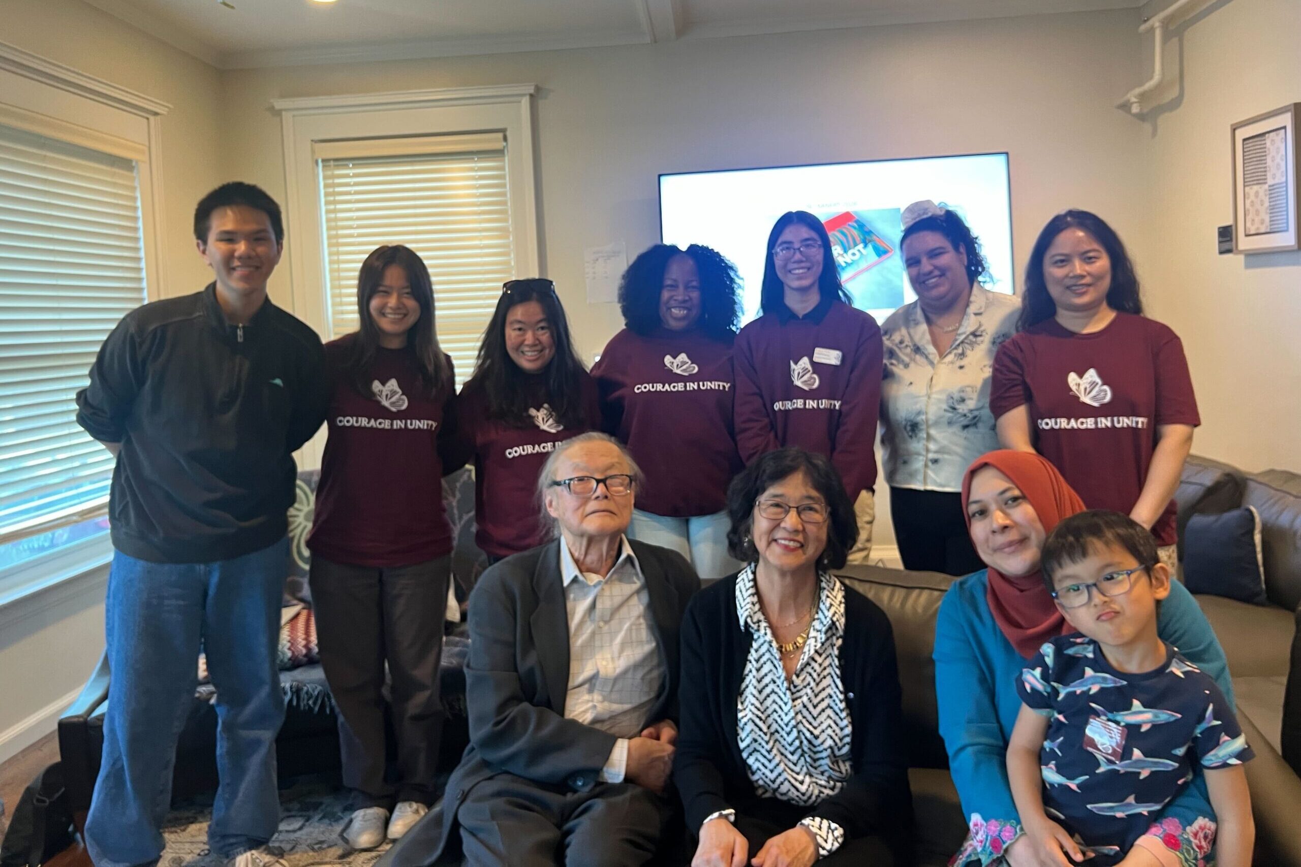 A group of young Asian American students stand behind professors sitting on a couch with a big TV and windows in the background.