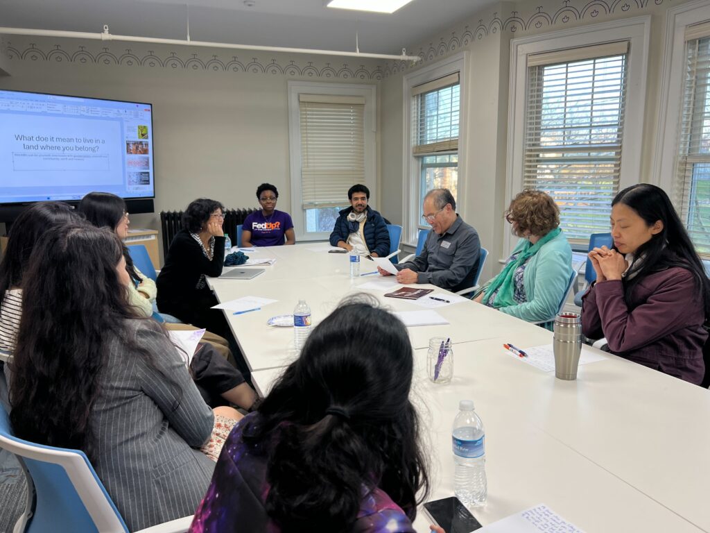 A female Asian American professor leads a group of students and faculty through a writing exercise on a long white table with sheets of paper and pens with windows and open blinds in the background.