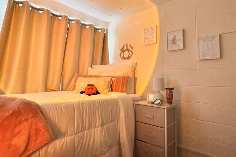 A cozy dorm room complete with beige curtains, a white duvet on a twin bed where an orange flow blanket and a stuffed Otto the Orange sit.