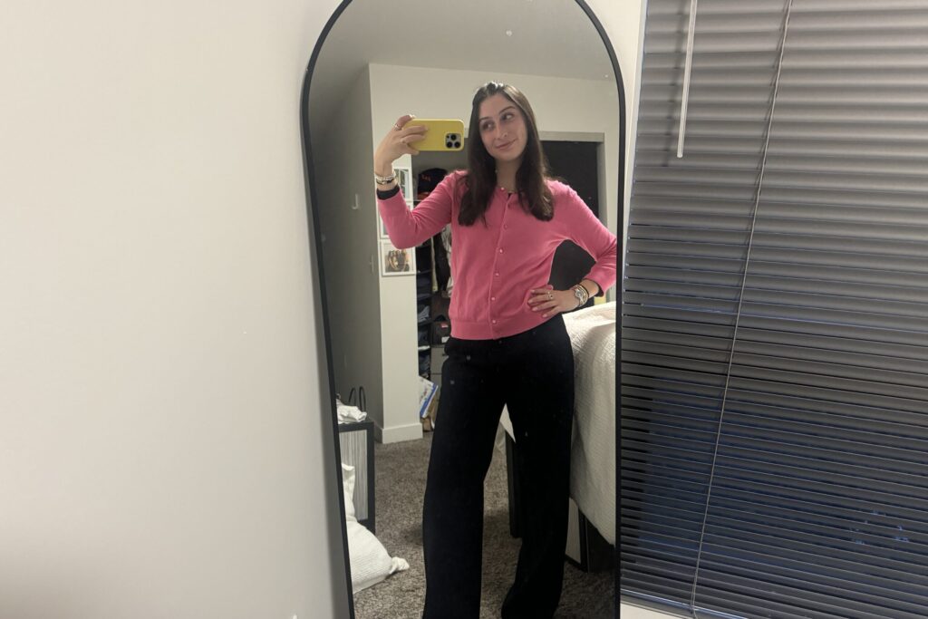 A woman takes a selfie in a floor-length mirror wearing a hot pink cardigan and black slacks.