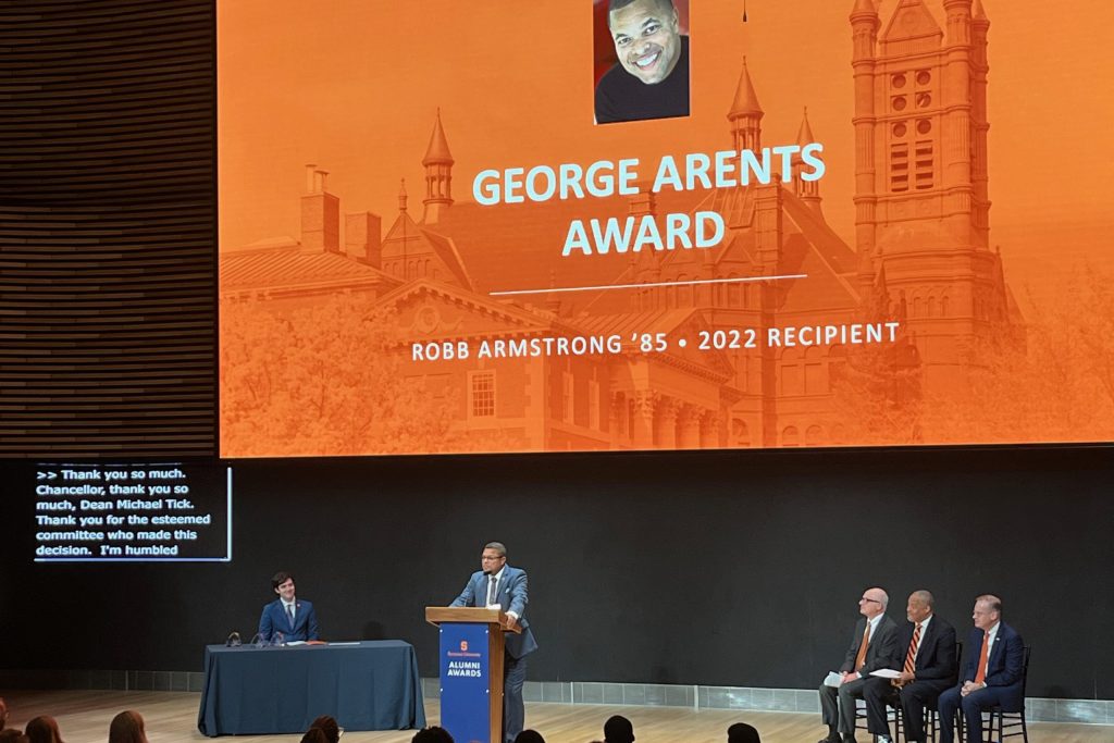 Syracuse Class of '85 alum Robb Armstrong stands at a podium in front of a darkened auditorium, with an orange screen behind him that says