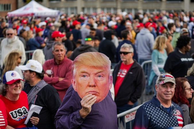 A Trump rally attendee holds up a Trump mask in line outside of the Oncenter on Friday, Oct. 9, 2026.