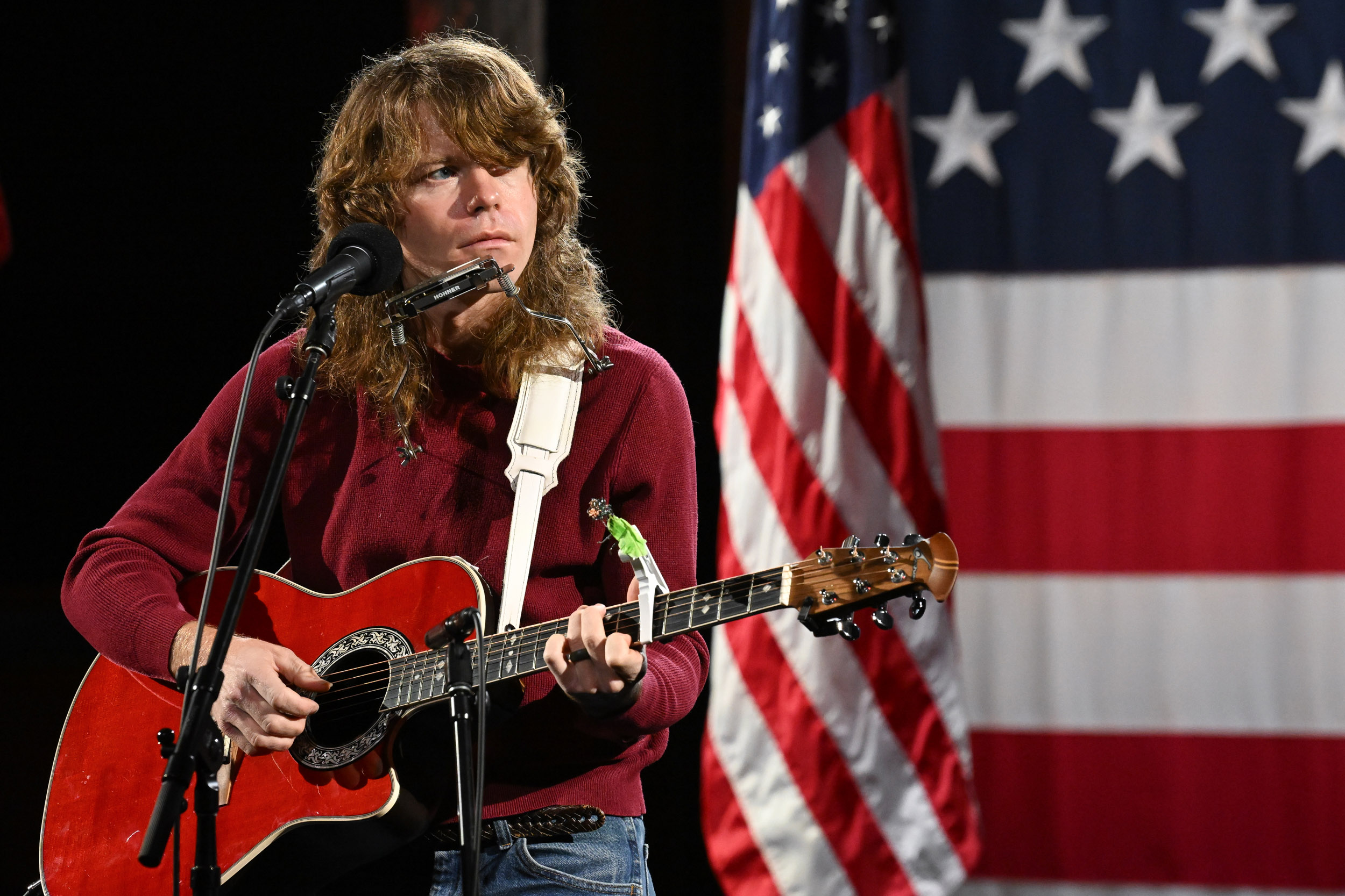 Man in red shirt with long brown hair strums a red guitar while sitting on a stool and American flag in the background.