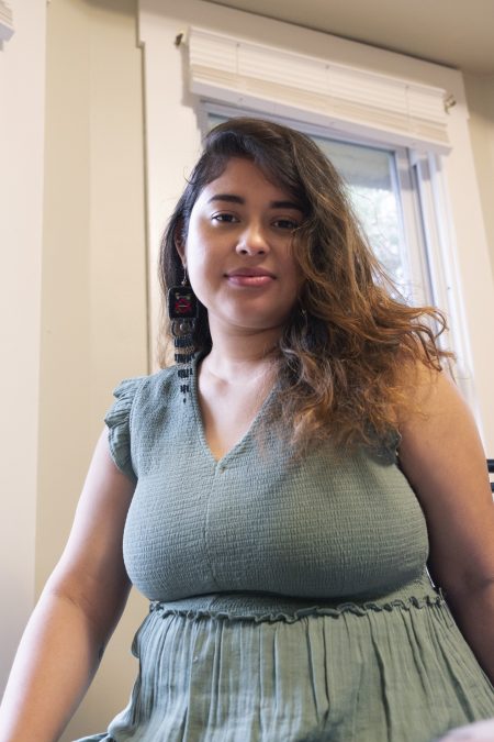 Angie Alberto sits upright in a softly lit room with windows just behind her head. Her face is lighter on her left side from the open windows, that reveal a soft smile. Her long dark brown hair cascades to the right in a side part. Long beaded earrings from Guatemala reach to her collarbone. She wears a green dress with gray undertones. It has a high v-neck cut with small ruflles along the shoulders. Her right arm is slightly forward, leaning on her knee for support, while the other rests at her side.