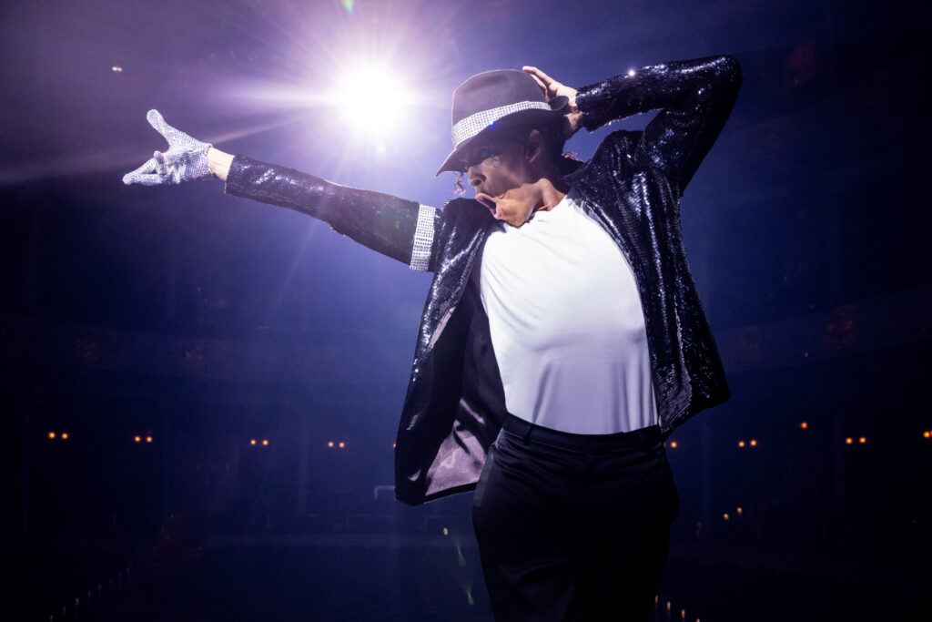 An actor playing Micheal Jackson stands alone on stage in his iconic black suit and sparkly glove.