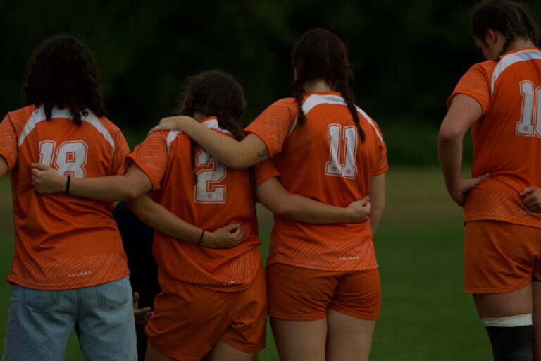 A group of girls in orange jerseys on Syracuse University's womens rugby team stand with their arms around each other.