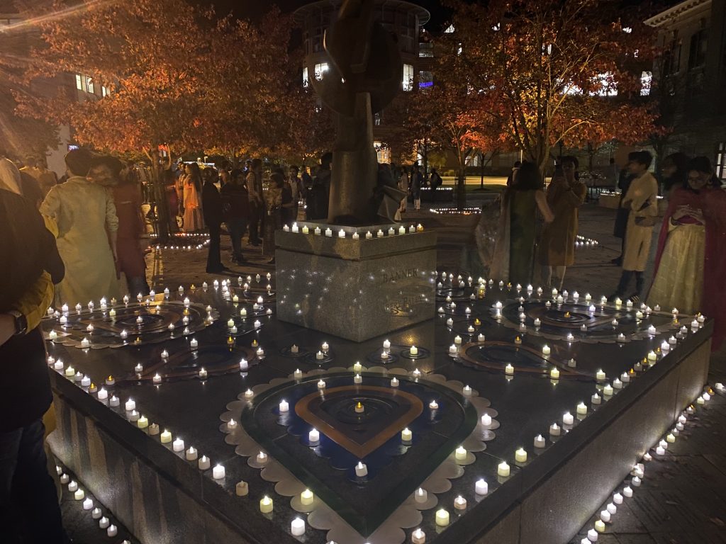 Colorful cut outs make up pretty patterns and shapes on the elevated base of a statue. Candles line the base of the statue as well and surrounding the rangoli designs. People are scattered around the statue, talking to each other and observing the lights and colorful display.