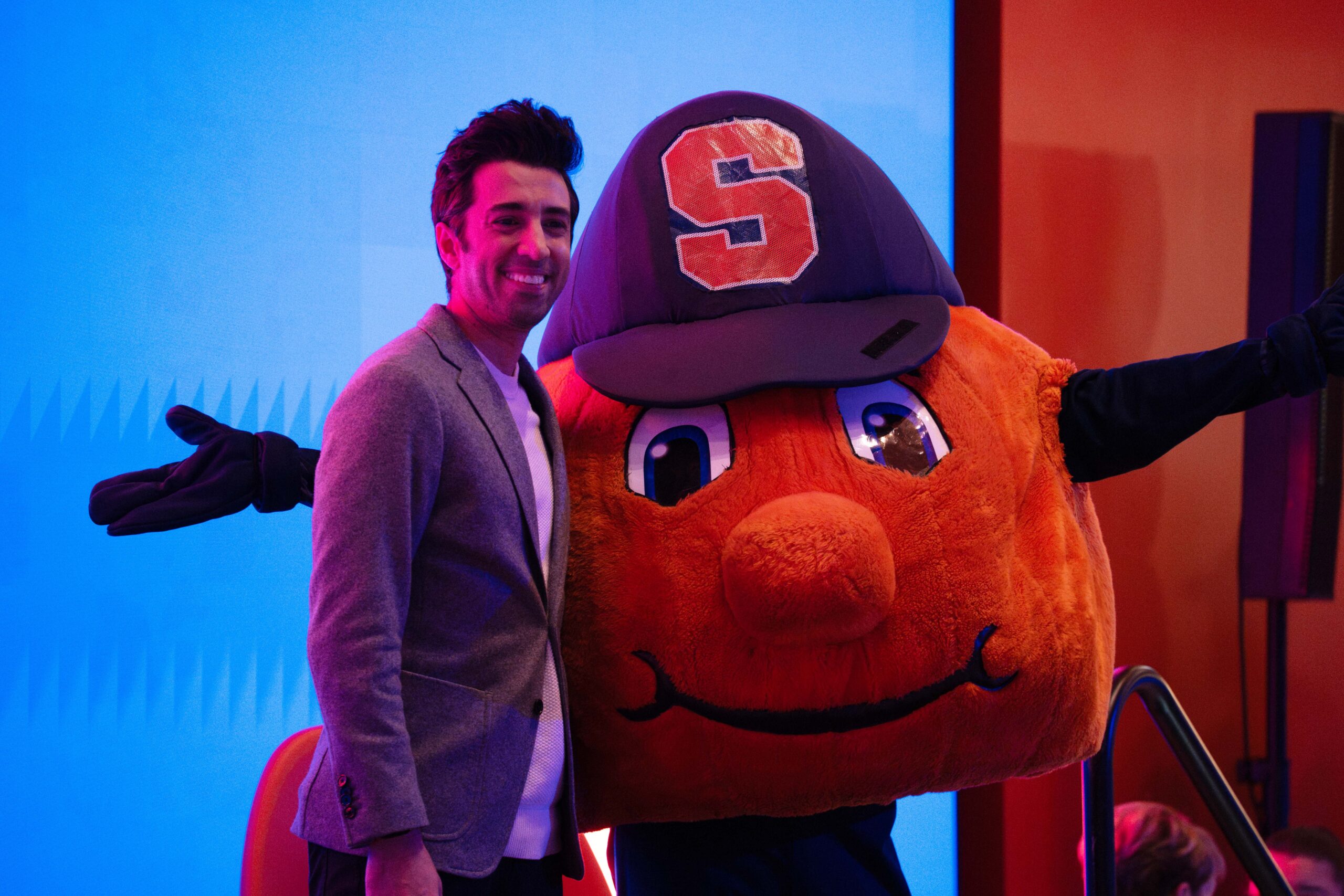 Man, Jon Yousheai, stands with Otto the Orange mascot on a stage in front of a blue screen.