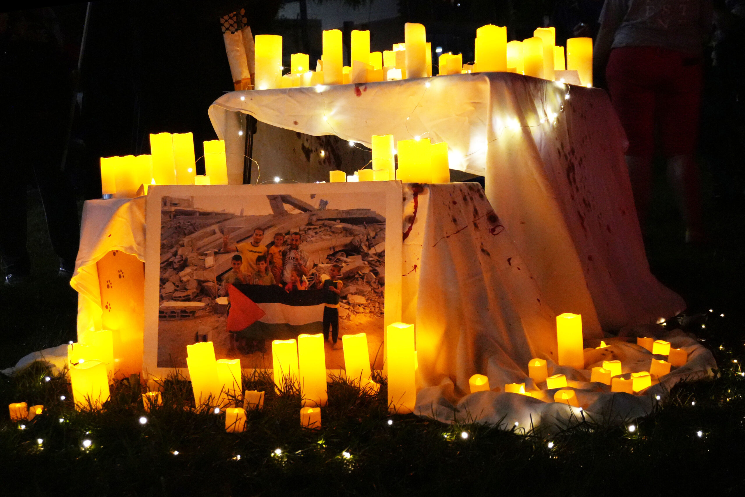 Candles surround a picture of a Palestinian family in Gaza, lit on a table with a fake bloody tablecloth at the Sumud Vigil for Gaza at the corner of Erie Boulevard and Genesee Street on Tuesday, October 7.