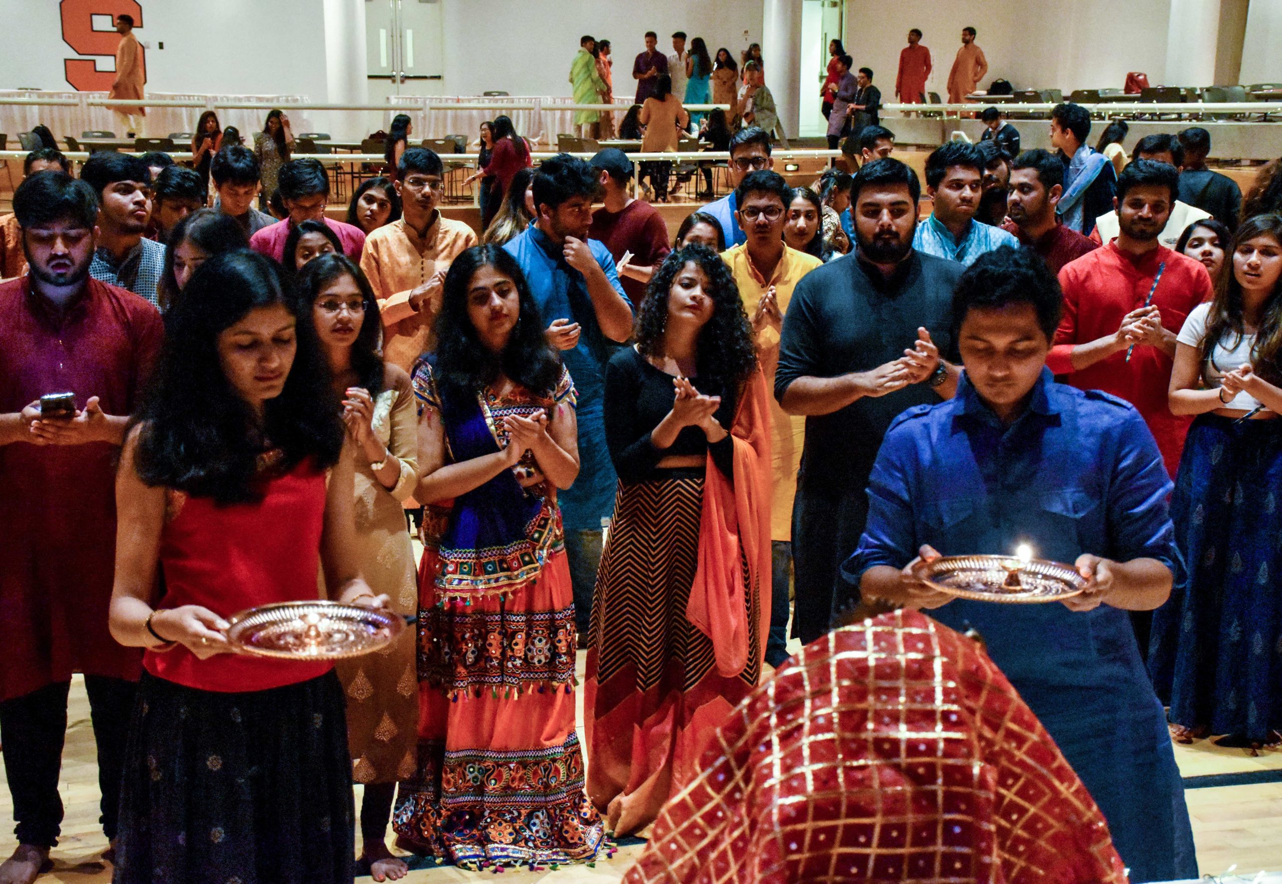 Group of students, brightly dressed, performing Aarti. With the front two totally engrossed in the worshipping with their eyes closed, holding aarti plates.