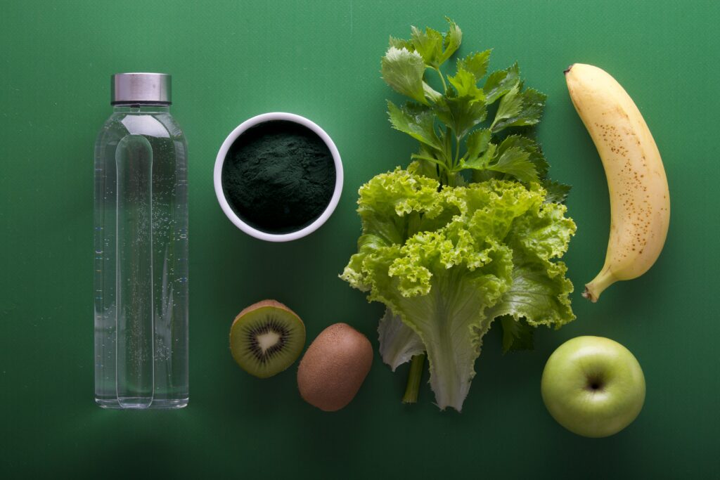 Various foods, including a water bottle, coffee mug, kiwi, lettuce, banana and apple sit against a green background.