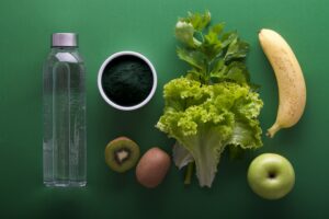 Various foods, including a water bottle, coffee mug, kiwi, lettuce, banana and apple sit against a green background.