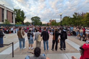 A shot from Syracuse Unversity campus taken on the steps of the library shows men lined up in front of a crowd of almost 100 people.