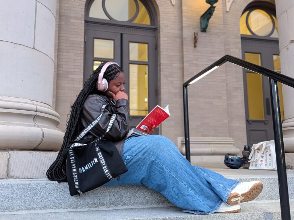 A women sits on the steps of a library in a leather jacket, with a book in hand and wearing over the ear headphones.