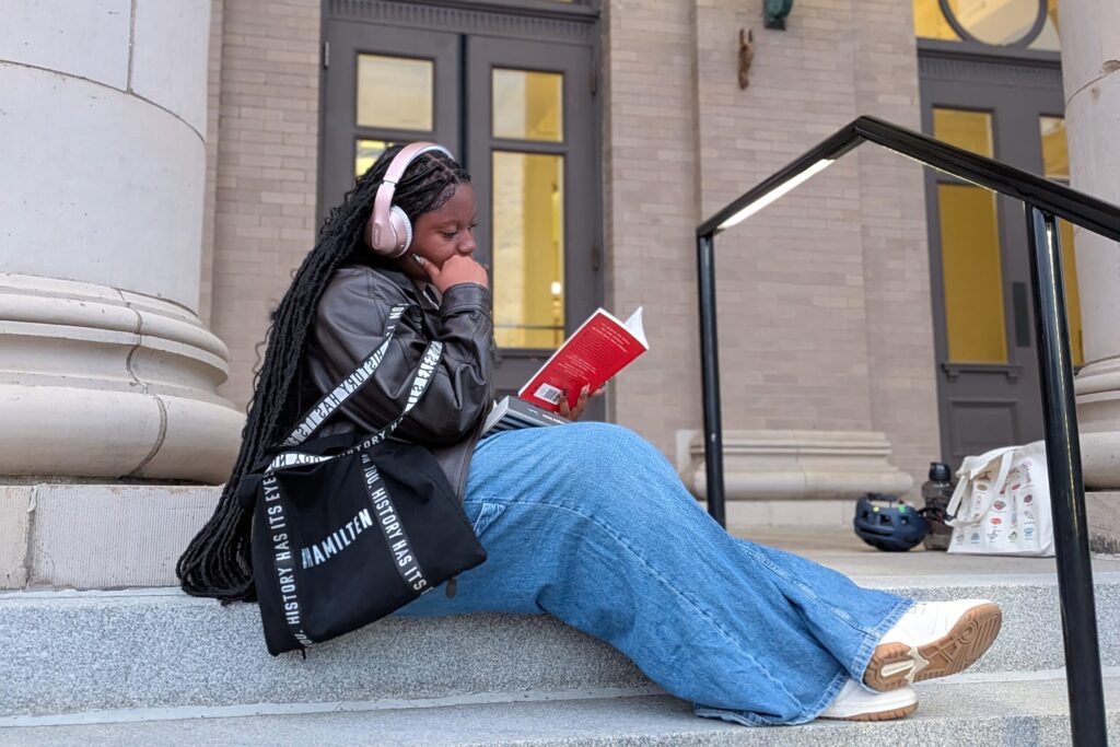 A women sits on the steps of a library in a leather jacket, with a book in hand and wearing over the ear headphones.