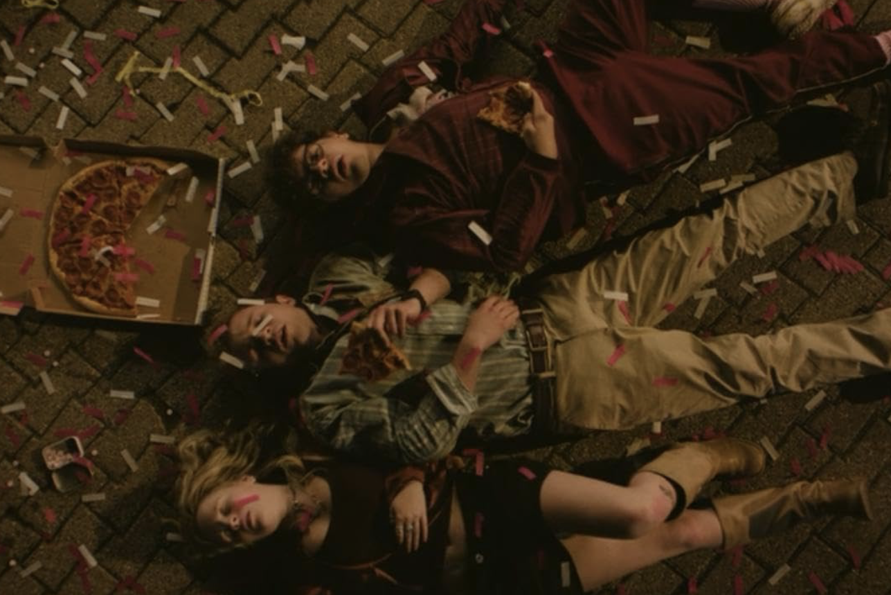 In "Pizza Movie" the three main characters lay on the ground covered in confetti, holding onto slices of pizza.