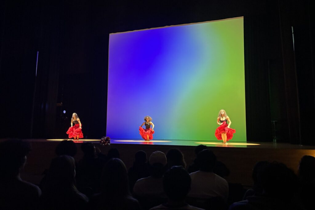 In front of a blue and green background, three dancers in long red skirts perform onstage.