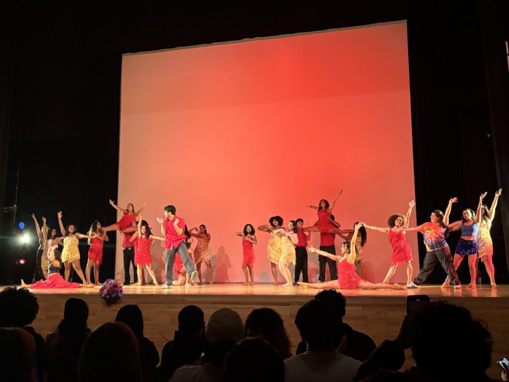 In front of a red background, a multitude of dancers perform onstage.