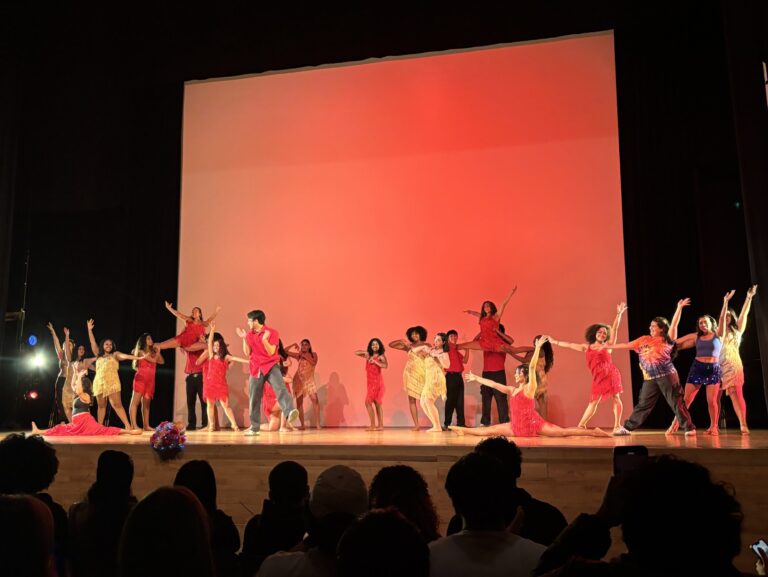 In front of a red background, a multitude of dancers perform onstage.
