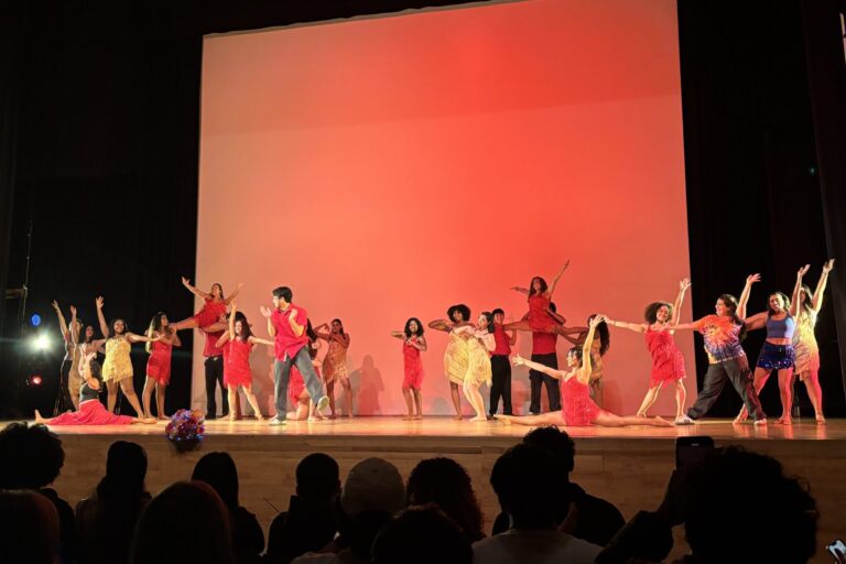 In front of a red background, a multitude of dancers perform onstage.