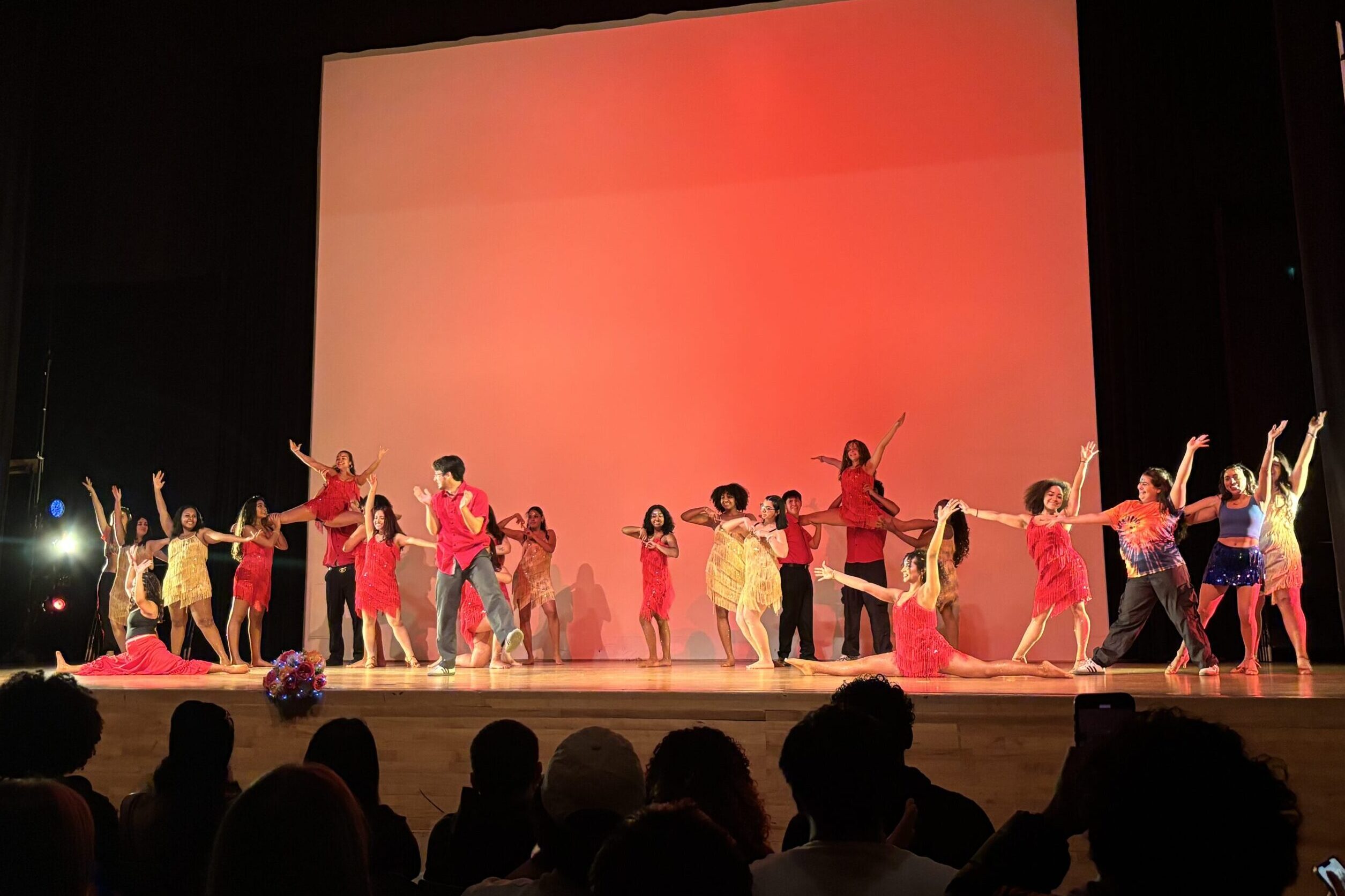 In front of a red background, a multitude of dancers perform onstage.