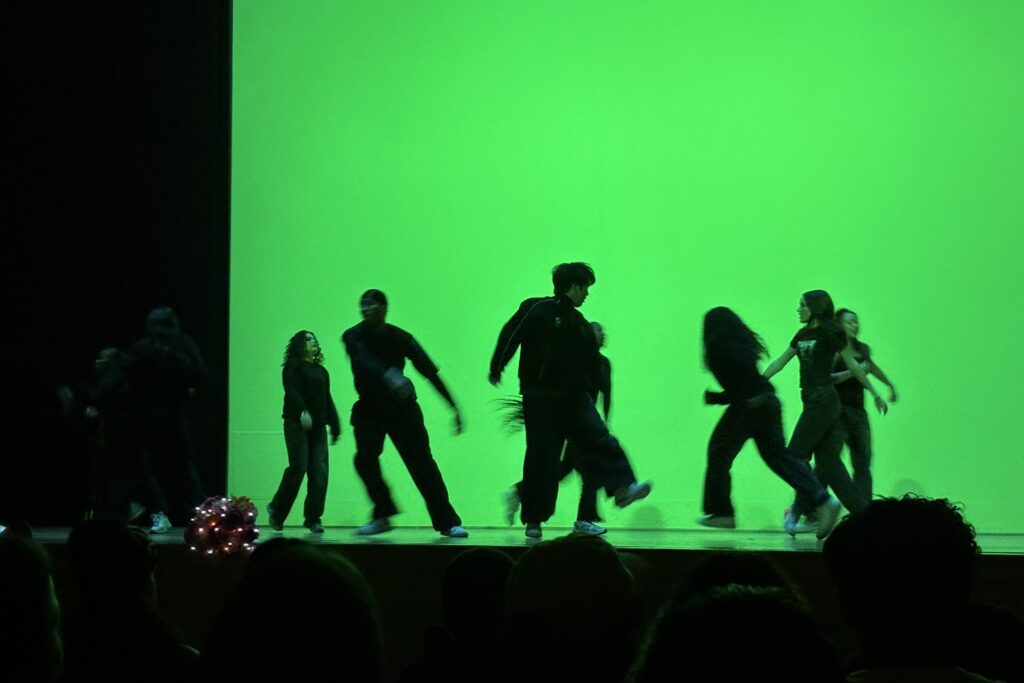 In front of a green background, dancers in all black perform onstage.