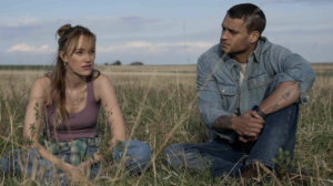 In "Reminders of Him" a woman dressed in a purple tank top sits in the grass next to a man dressed in denim.