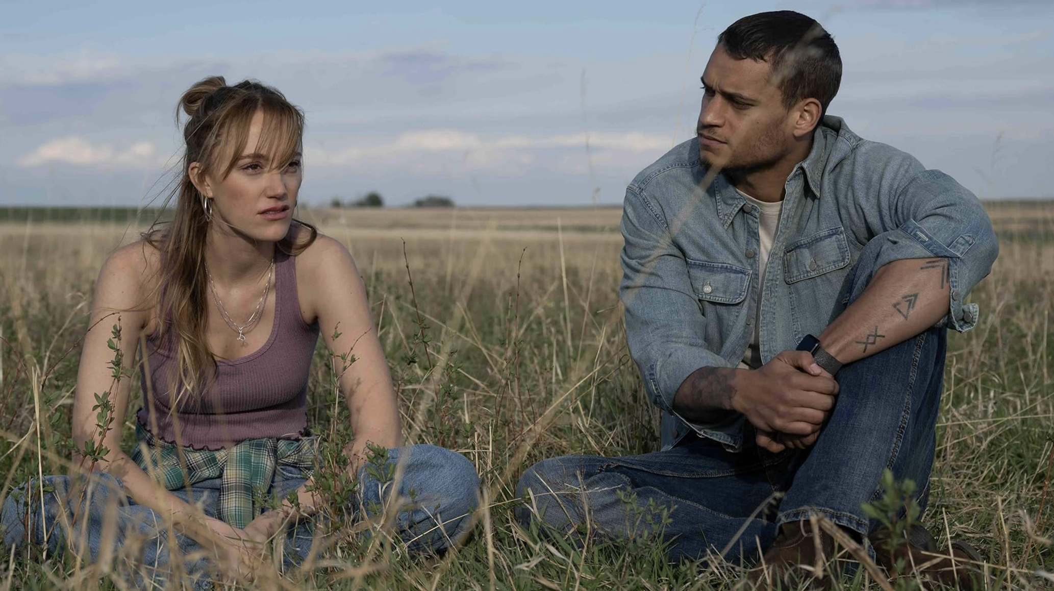 In "Reminders of Him" a woman dressed in a purple tank top sits in the grass next to a man dressed in denim.