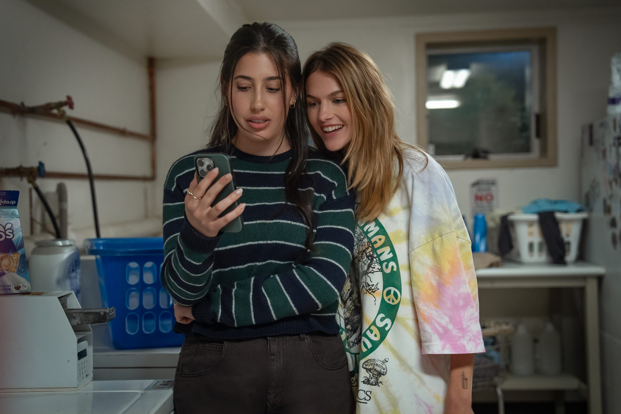 In "Roommates" Sadie Sandler and Chloe East stand together looking at a phone.